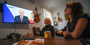 Leute sitzen an einem Tisch mit Essen und schauen auf einen Bildschirm mit Putin