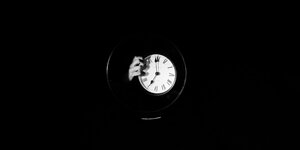 Aus einem schwarzen Hintergrund kommt eine Hand und hält eine Uhr, sie zeigt die Uhrzeit fünf Uhr morgens