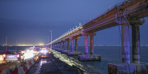 Nächtliche Szene auf einer Brücke übers Meer, Fahrzeuge sind unterwegs, im Vordergrund sind Beschädigungen zu sehen