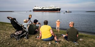 Menschen sitzen am Wasser auf einer Wiese.