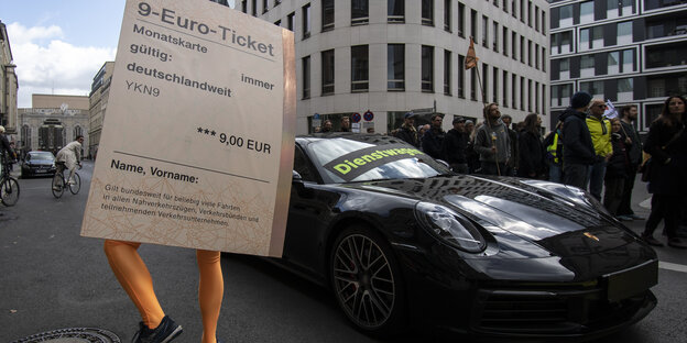 Ein Mensch hinter einem riesigen 9-Euro-Ticket neben einem Porsche