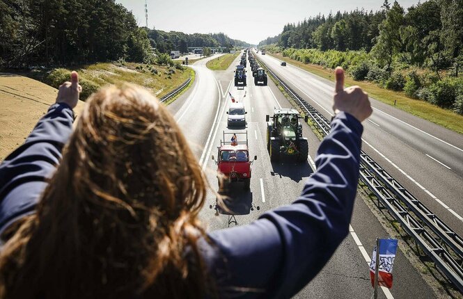 Eine Frau steht auf einer Brücke über einer Autbahn. Unter ihr fährt eine Kolonne aus Traktoren durch. Sie zeigt ihnen zwei Daumen hoch, um Zustimmung auszudrücken