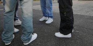 Vier Menschen stehen auf einem Schulhof in Jeans und weißen Sneakern im Kreis, die Oberkörper sind nicht zu sehen