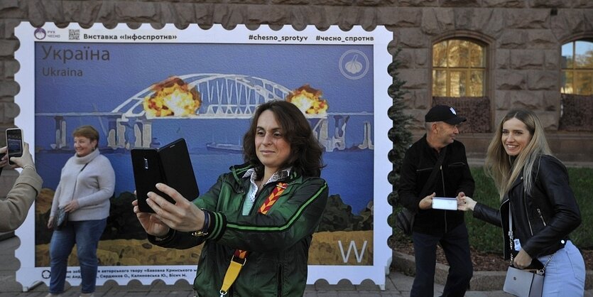 Eine Frau fotografiert sich selbst vor einer überdimensional großen briefmarke mit dem Motive der brennenden Krim-Brücke