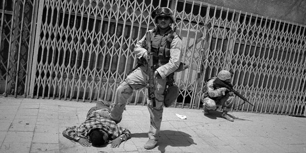 Ein Mann liegt mit dem Gesicht nach unten auf der Straße, ein US-Soldat mit Maschinengewehr steht mit einem Bein auf seinem Rücken