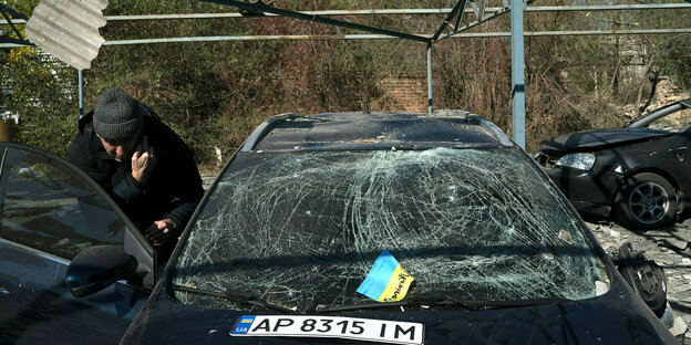 Ein Mann steht zwischen seinem Auto und der geöffneten Beifahrertür und telefoniert. Auf der zerborstenen Windschutzscheibe liegt eine kleine ukrainische Flagge