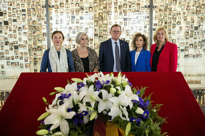 Bodo Ramelow steht in einer gruppe von menschen, hinter ihm ist eine Fotowand mit vielen Portraits, vor ihm liegt ein Blumenkranz