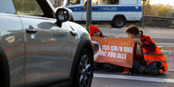 Zwei junge Frauen sitzen auf der Straße mti einem Plakat: 100 km/h und 9 € für alle - vor ihnen steht ein Auto