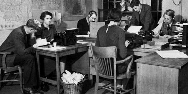 historische Schwarz-Weiß-Aufnahme: Frauen udn Männer an SChreibtischen in einem Redaktionsbüro, Weltkarte im Hinterrund, einige tragen Kopfhörer, ein Papierkorb quillt über