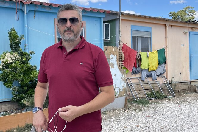 ein Mann mit Sonnenbrille und rotem Poloshirt steht vor bunten Hütten