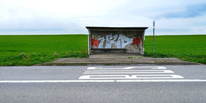 Eine Bushaltestelle steht einsam in der Landschaft, auf dem Asphalt steht groß das Wort "Bus"