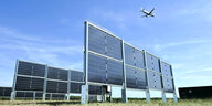 Solarpanele stehen aufrecht auf einer Wiese, darüber fliegt ein Flugzeug