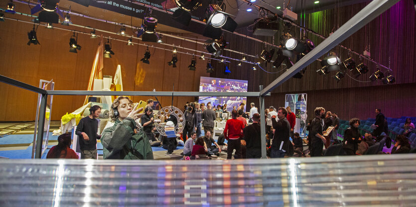 Installation im Haus der Kulturen der Welt, viele Menschen stehen auf der Bühne, im Hintergrund ein Berg aus Teppichbodenrollen. Im Vordergrund fotografiert eine Frau eine Trophäe, die an einem Gestell hängt