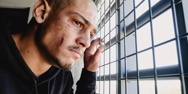 Ein Mann steht an einem vergitterten Fenster und blickt nach draußen. Seine Hand greift ans Gitter