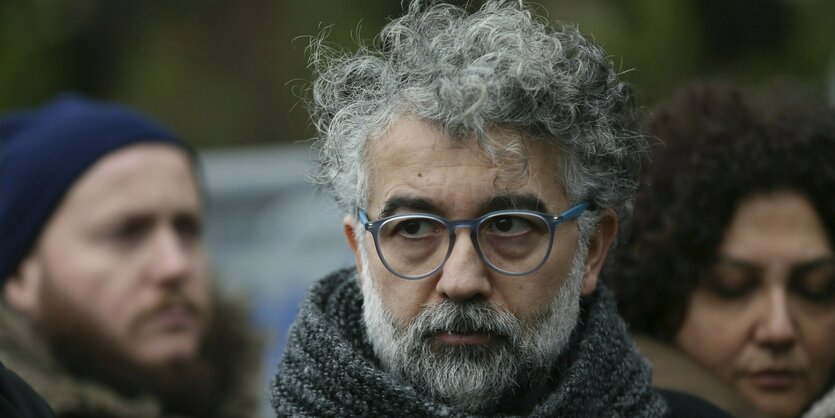 Erol Onderoglu im Portät. Er hat lockiges grau-schwarzes Haar, einen Bart in der gleichen Farbe und eine runde Brille. Hinter ihm zwei Personen.