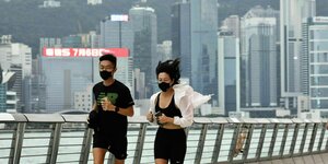 Ein Paar joggt über eine Brücke, im Hintergrund die Skyline von Hongkong. Beide tragen eine schwarze FFP2 Maske