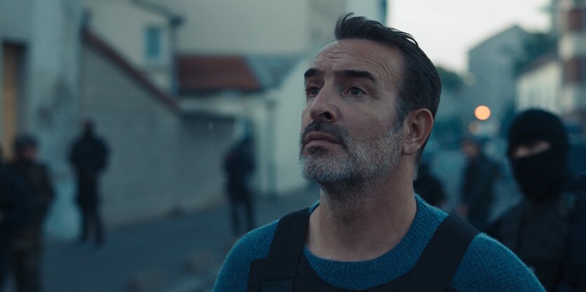 Fred (Jean Dujardin) steht mit uniformierten Kollegen von der Antiterroreinheit der französischen Polizei auf einer Straße.