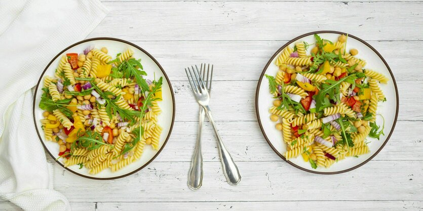 Zwei Teller mit Nudeln, Gemüse und Ruccola stehen auf einem hellen Holztisch