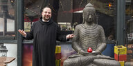 Der Benediktinermönche steht mit Kutte neben einer lebensgroßen Buddhafigur
