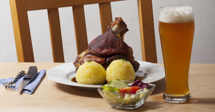 Ein Teller mit Schweinshaxe und Klößen. Ein Weizenbier rechts vom Teller. Besteck links vom Teller. Davor ein kleiner gemischter Salat.