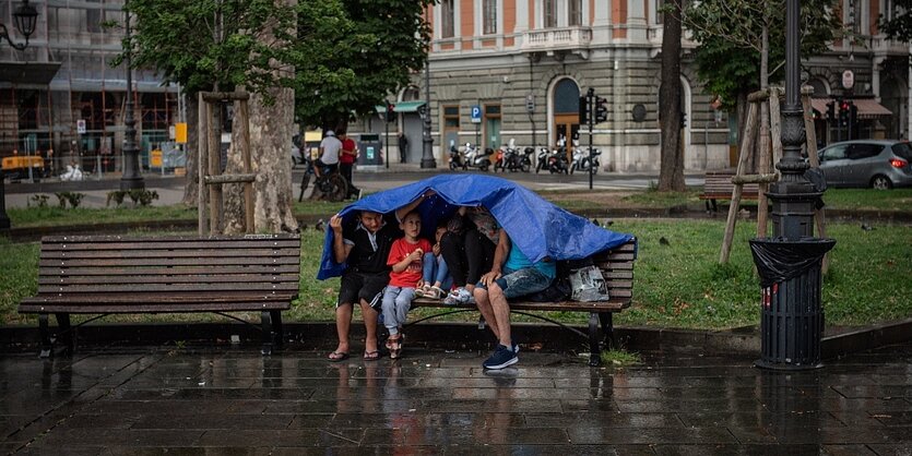 Eine Familie sitz auf einer Parkbank und schützt sich mit einer blauen Plane vor dem Regen