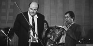 Schwarz-weiße Aufnahme: Der Musiker Manfred Krug steht mit Anzug und Krawatte auf der Bühne an einem Mikrophonständer, der auf die Klarinette gerichtet ist, in die der Komponist Günther Fischer links neben ihm hinein bläst