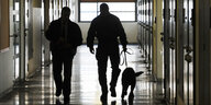 Zwei Männer in Uniform gehen mit einem Hund an der Leine über einen Zellenflur
