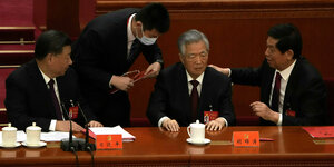 Zwei Saaldiener wenden sich dem sitzenden Hu Jintao zu, der versteinert wirkt. Auch der neben ihm sitzende Xi wendet ihm den Kopf zu