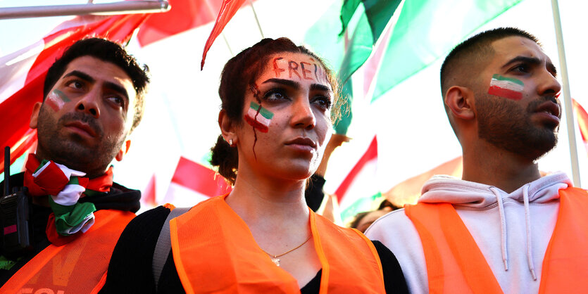 Zwei Männer rahmen eine Frau ein, sie stehen vor einem Meer iranischer Flaggen, schauen ernst und haben selbst ihr Gesicht mit der iranischen Fahne bemalt