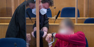 Ein Mann im roten Pullover auf der Anklagebank ist verpixelt, er hat Handschellen und begrüßt seinen Anwalt mit einem coronamäßigen Faustknöchelgruß