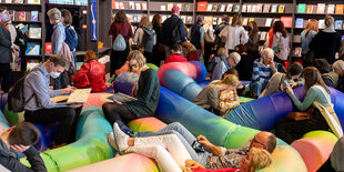 Viele Menschen liegen auf bequemen Sesseln, im Hintergrund laufen Besucher der Buchmesse an Bücherständen vorbei