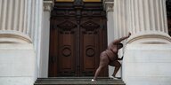 Eine Schwarze Frau steht nackt an der Säule eines Gerichtsgebäudes und sieht so aus, als schiebe sie die Säule zur Seite