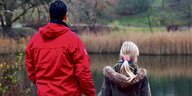 Mann in roter Jacke und Kind mit blondem Pferdeschwarz schauen auf einen Teich