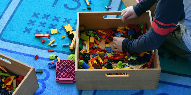 Ein Kind wühlt in einer Kiste mit Bausteinen auf einem blauen Teppich