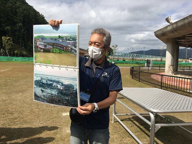 Ein Mann mit Mundschutzmaske steht vor einer zerstörten grundschule und hält zwei Fotos, die die Folgen des Tsunamis zeigen, in der Hand