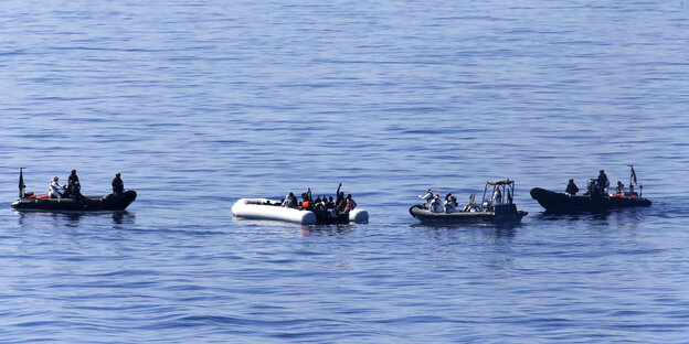 3 dunkle kleine Motoboote mit Soldaten umringen ein kleines Schlauchboot mit vielen Menschen