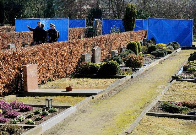 Gräber mit Hecken, dahinter blaue Plastikplanen um die Exhumierung abzuschirmen. Vier Polizisten stehen Wache