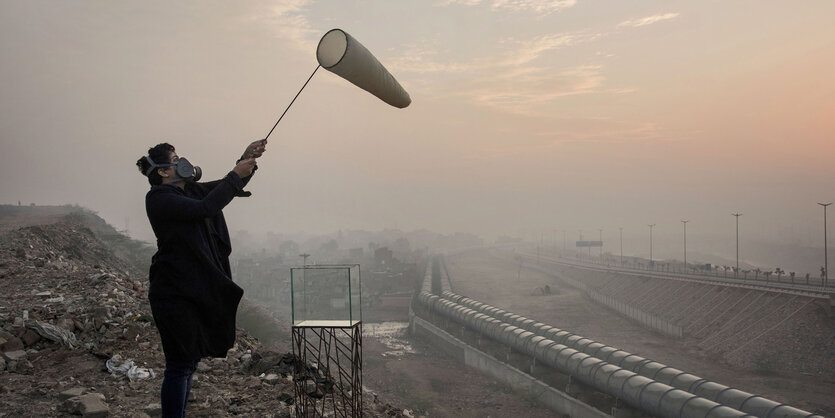 Eine Frau mit Gasmaske hält einen Stoffsack in die Luft, im HIntergrund eine Stadt im Smog.