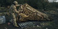 Ein gemusterter Teppich wird von einem Sodlaten aufgedeckt, darunter versteckte Munition
