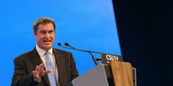 Markus Söder steht an einem Pult mit Mikro und spricht vor einem blauen Hintergrund