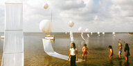 Ballons schweben über dem Meer, Segel hängen daran, Kinder waten durchs Wasser