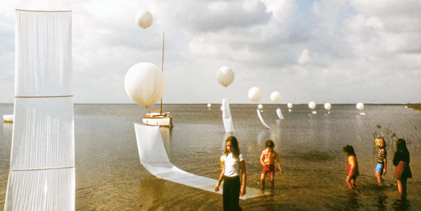 Ballons schweben über dem Meer, Segel hängen daran, Kinder waten durchs Wasser