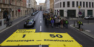 gelbe Bahnen mit Aufschrift "Herr Lindner 1 Dienstwagen oder 50 Klimatickets" bedecken die Straße ovr der FDP-Parteizentrale, ein Demozug nähert sich
