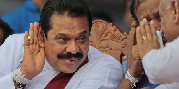 Mahinda Rajapaksa hält sich eine Hand hinter das Ohr