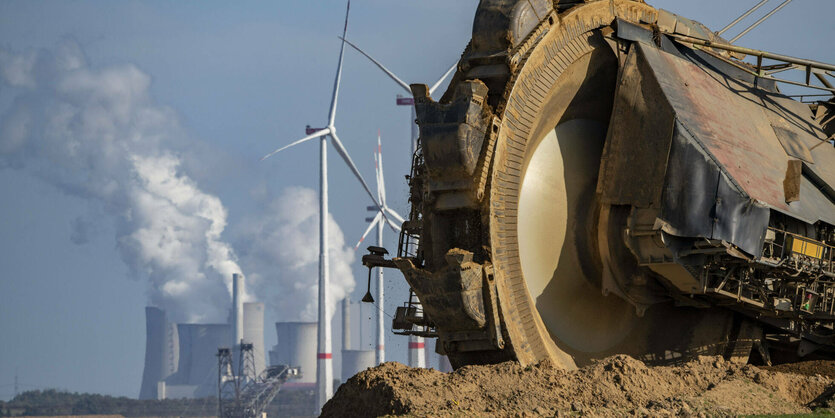 Riesiges Baggerrad vor blauem Himmel, dahinter dampfendes Kraftwerk und mehrere Windräder