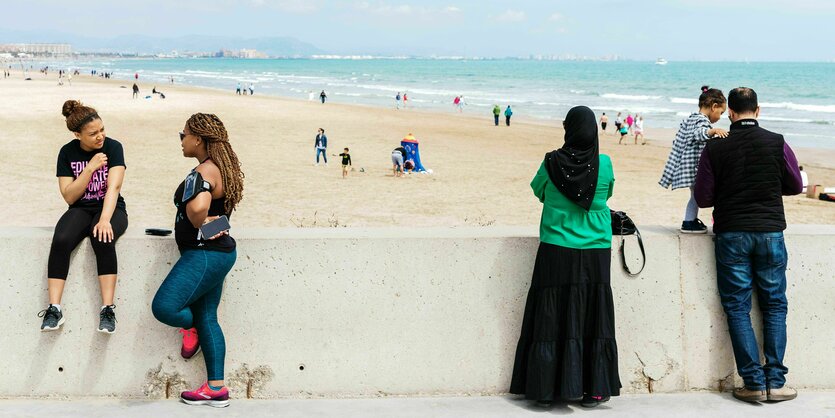 An einem Strand von Valencia stehen Menschen an einer hüfthohen Mauer, darunter eine Frau mit Kopftuch