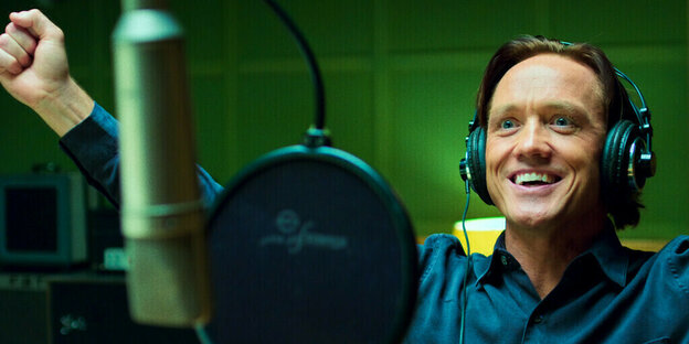 Ein Mann mit Kopfhörern sitzt in einem Studio und reißt lachend die Arme hoch