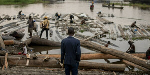 Das Foto zeigt einen Mann im Anzug, der das Ankommen von Baumstämmen in einem kleinen Hafen am Kongo beobachtet