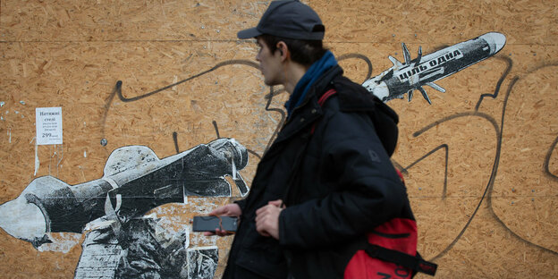 A young man in a black jacket with a black cap and a red backpack is walking along in front of a wooden shack.  On the stall is street art: a human with a weapon resembling a bazooka.