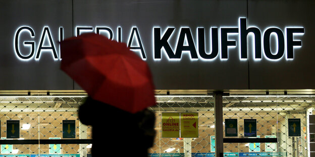 Das Bild zeigt einen Menschen mit rotem Regenschirm, der an einer geschlossenen Galeria-Kaufhof-Filiale vorbeigeht.